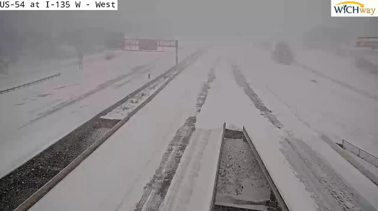 Kansas DOT traffic camera looking west on US-54 at I-135 during heavy snowfall. The highway is fully snow-covered with two dark wheel tracks worn through to the pavement.
